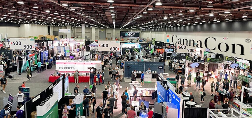A large indoor convention hall filled with booths, displays, and people attending CannaCon Midwest Lansing 2025, a cannabis industry expo. Signs with numbers mark different sections as various vendors interact with visitors.