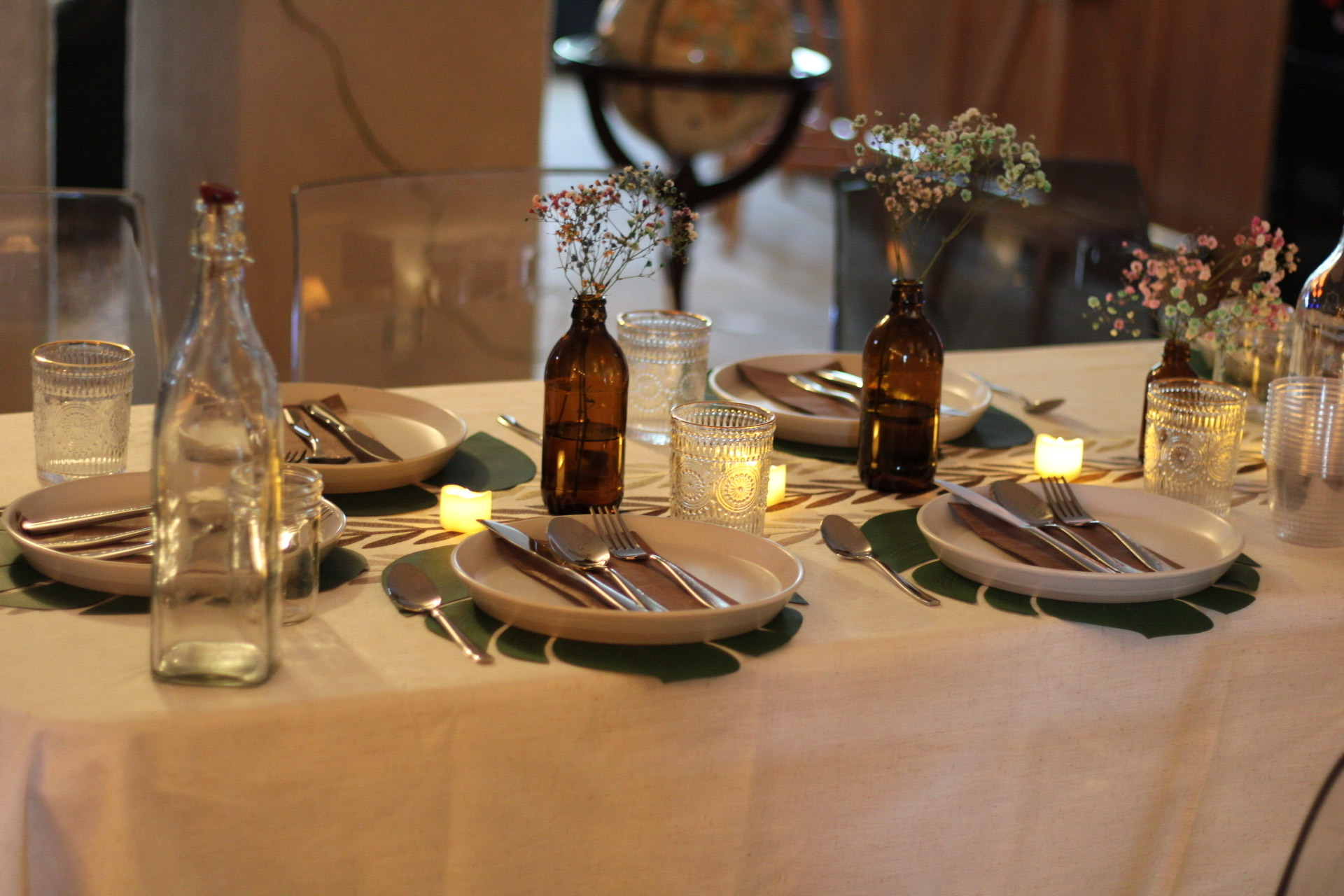 A table set for four evokes a Faded Feast, with white plates, cutlery, clear glasses, brown bottles holding baby's breath flowers, small candles, and a white tablecloth accented by green leaf placemats.